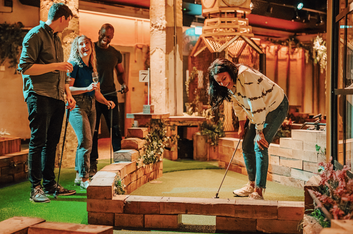 Two men and two women laugh with one another on the crazy golf course, as one of the women putts her ball.