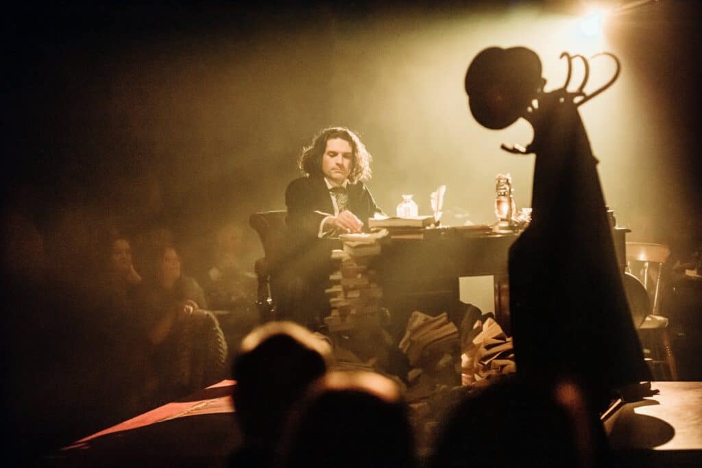 A man dressed as Charles Dickens sits at an old-fashioned desk, an audience faintly visible behind him.