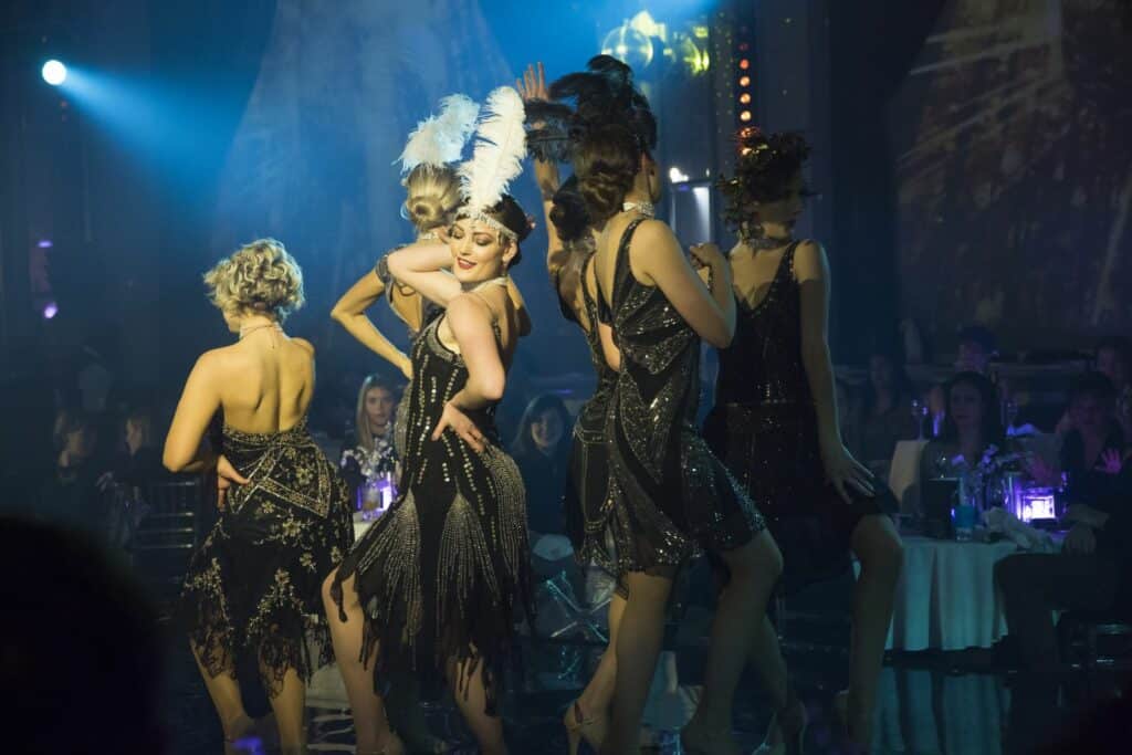 Cabaret performers in 1920s flapper dresses and feathered headpieces pose onstage.