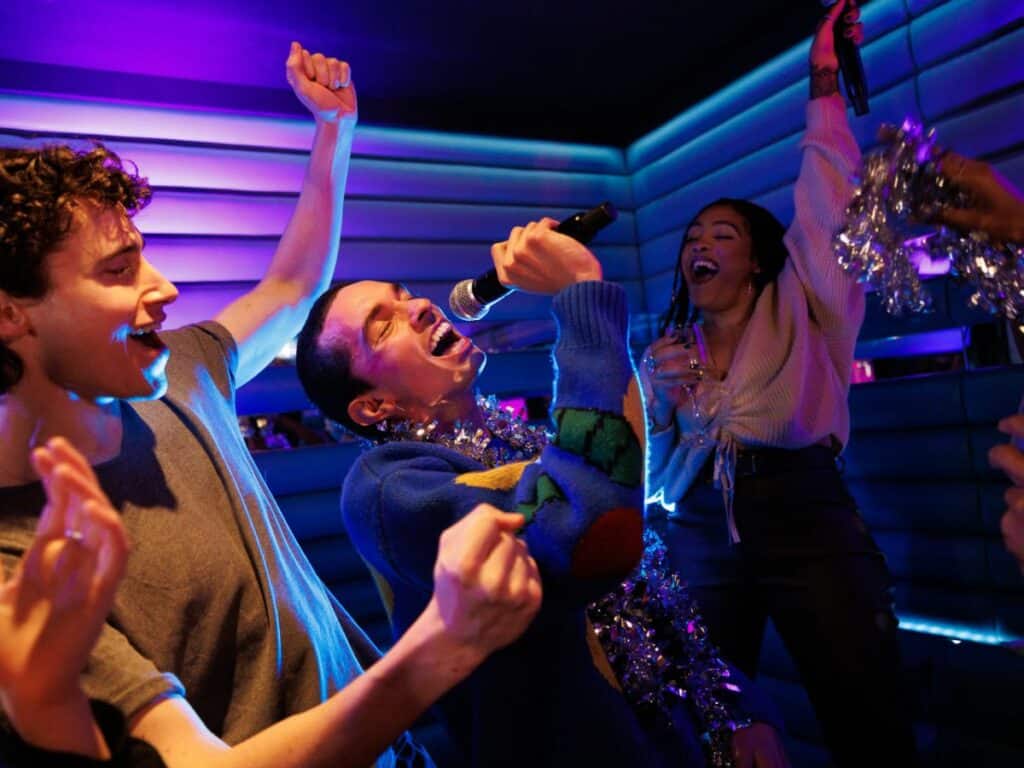 Three people sing and dance in a blue-lit karaoke room, the man in the middle leaning back to belt into the microphone.