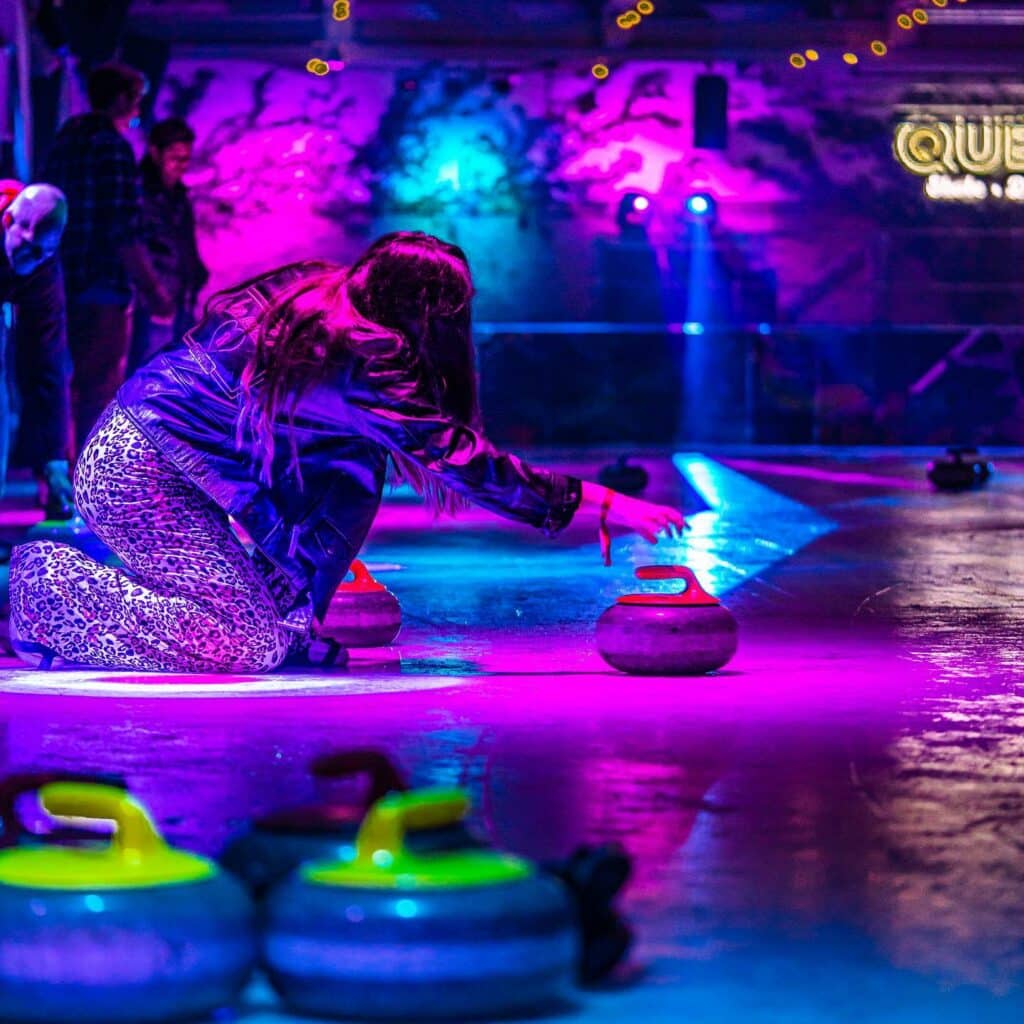 A young woman kneels on a blue-and-purple-lit curling court, attempting to curl on the ice.
