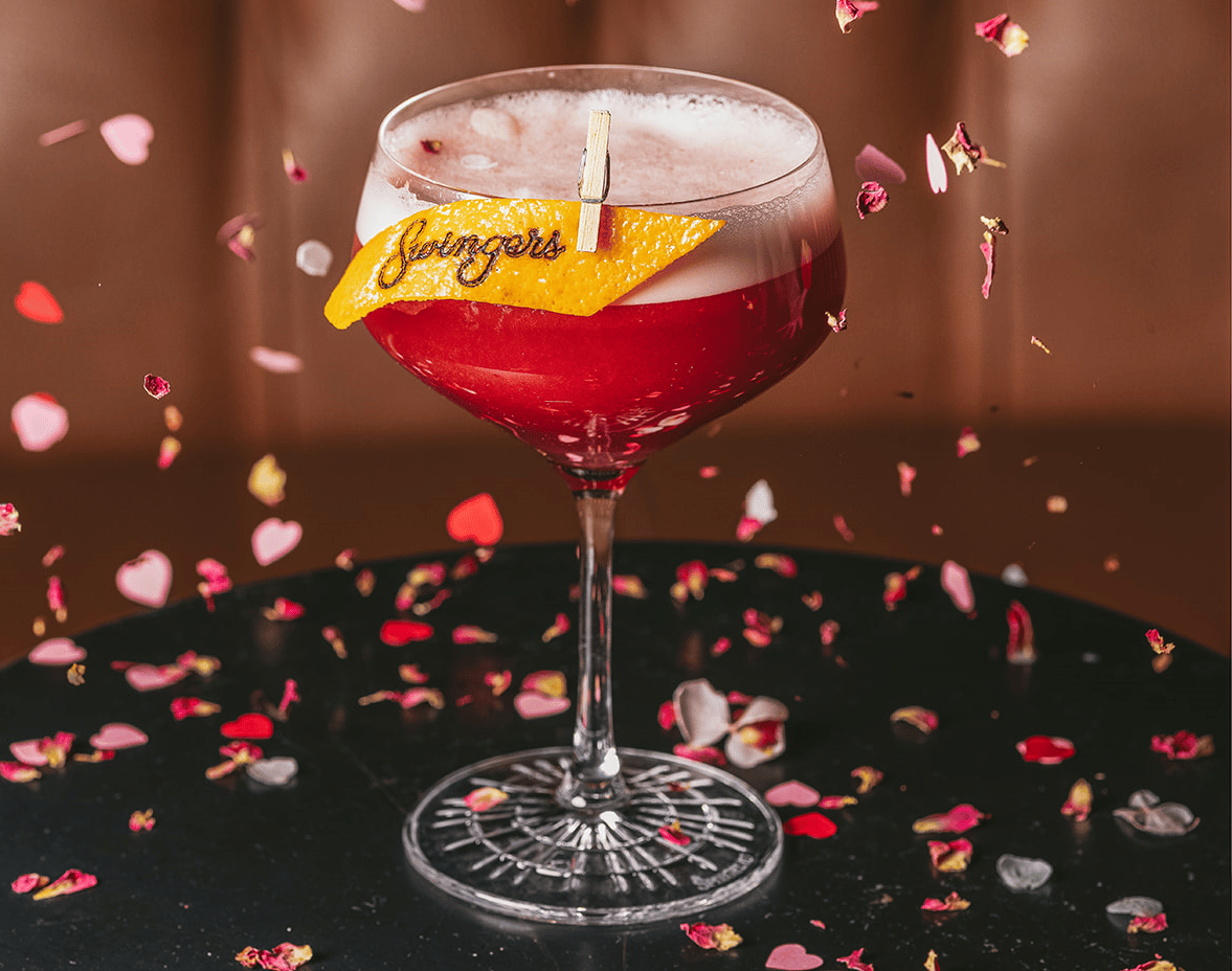 A berry-coloured cocktail garnished with an orange peel, surrounded by rose petals and heart-shaped confetti for Valentine's Day.