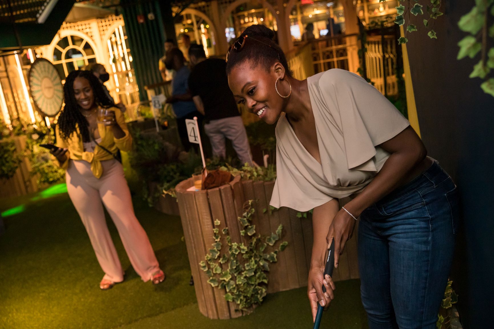 A young woman smiles, drink in hand, watching another young woman line up her shot on a crazy golf course.