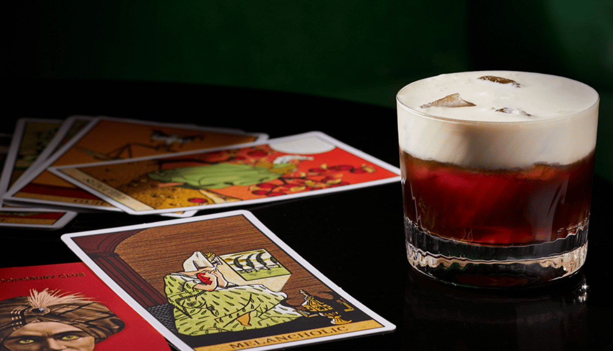 A dark liquor drink with a foamy finish sits beside a pile of tarot cards.