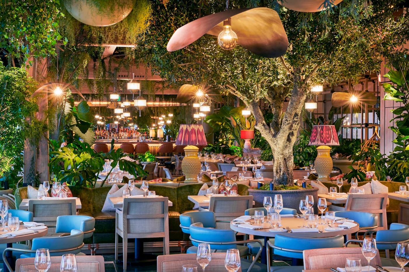 The interior of an upscale restaurant with leafy decorations, warm lights and pale pink-and-blue decor.