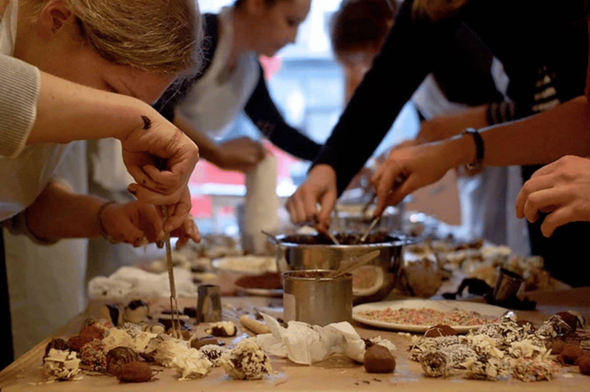 People crowd around a table making truffles with white chocolate shavings.