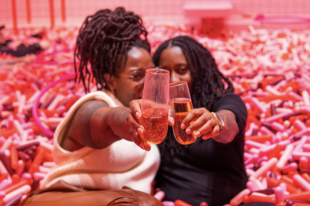A couple sits among pink-and-red plastic sprinkles, raising glasses of sparkling wine to the camera.