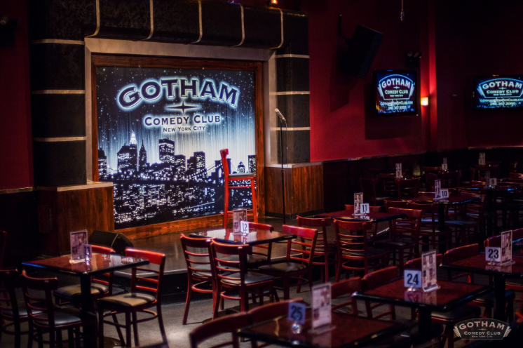 The interior of Gotham Comedy Club, with polished wooden tables and a stage mural featuring the city.