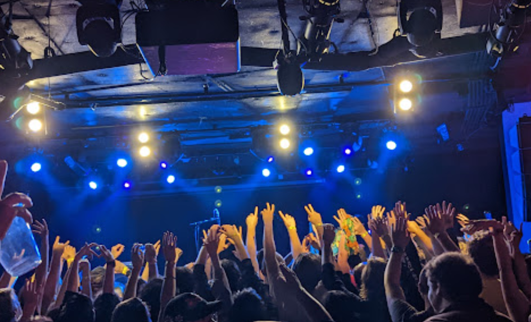 A blue-lit stage crowded by people with their hands in the air.