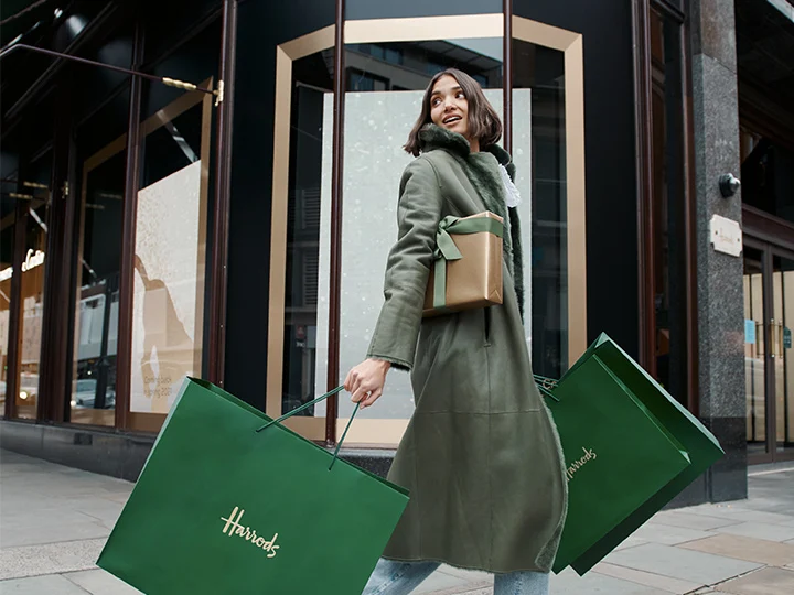 A young woman in a fur-lined coat emerges from Harrods, two large green shopping bags in her hands.