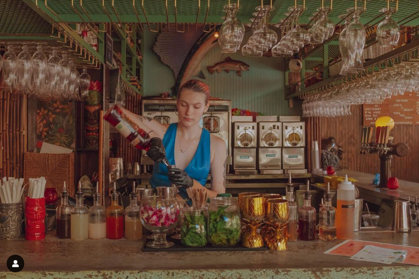 A bartender measures a drink behind a bartop covered in jars of garnishes and tiki bar decorations.