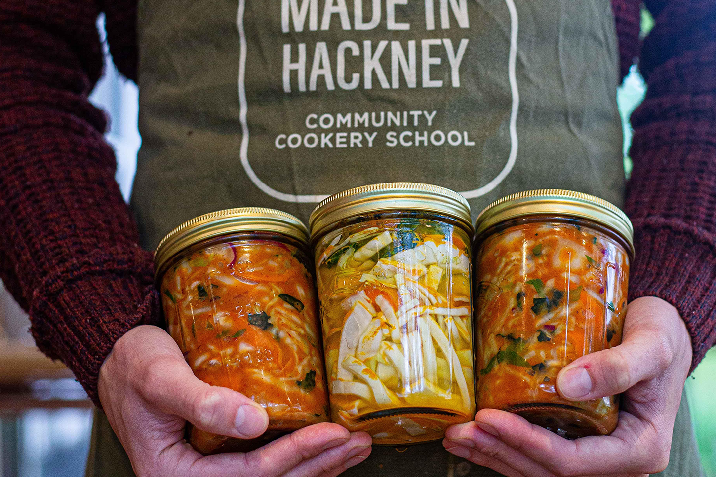 A man in a "Made in Hackney" apron holds three mason jars of vegan soup.