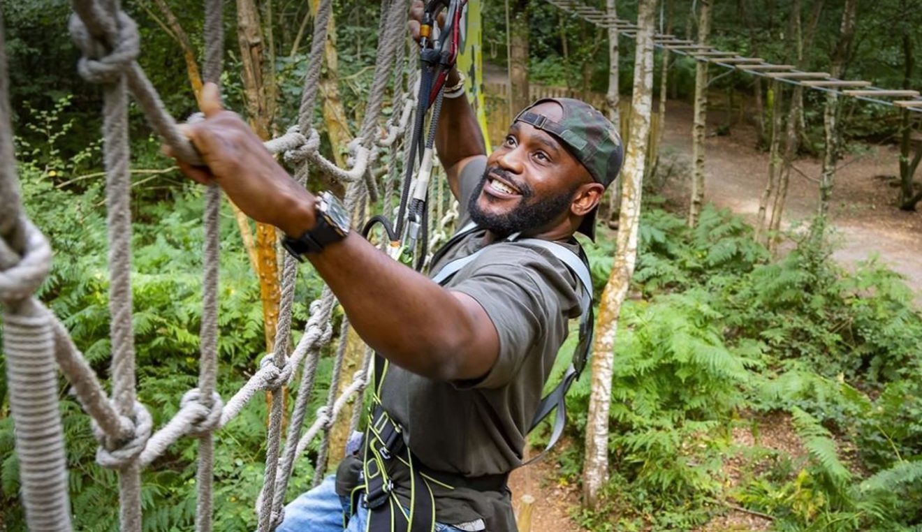 A man climbs a rope course through the trees, looking upwards to see where to place his hands.