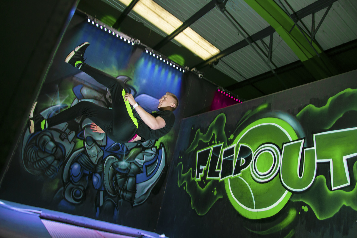 A man does a flip on a purple trampoline, the walls covered in decorative purple, green, and black graffiti.