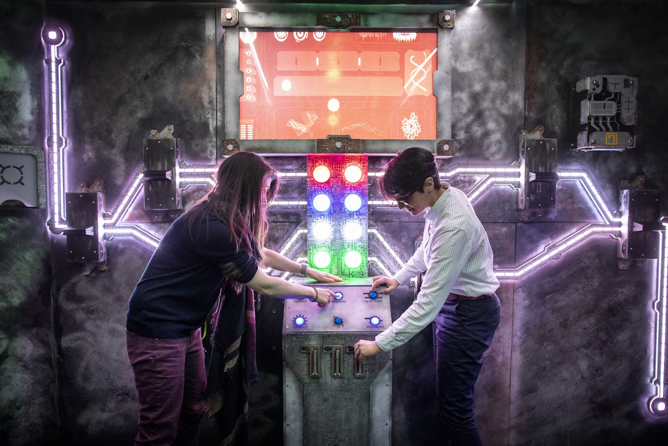 Two people operate a control panel in the centre of a futuristic neon-lit escape room.