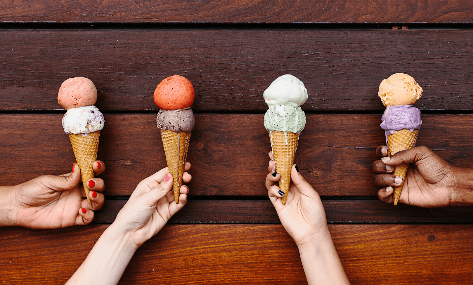 Four hands hold up four cones of ice cream, each with two scoops of different flavors.