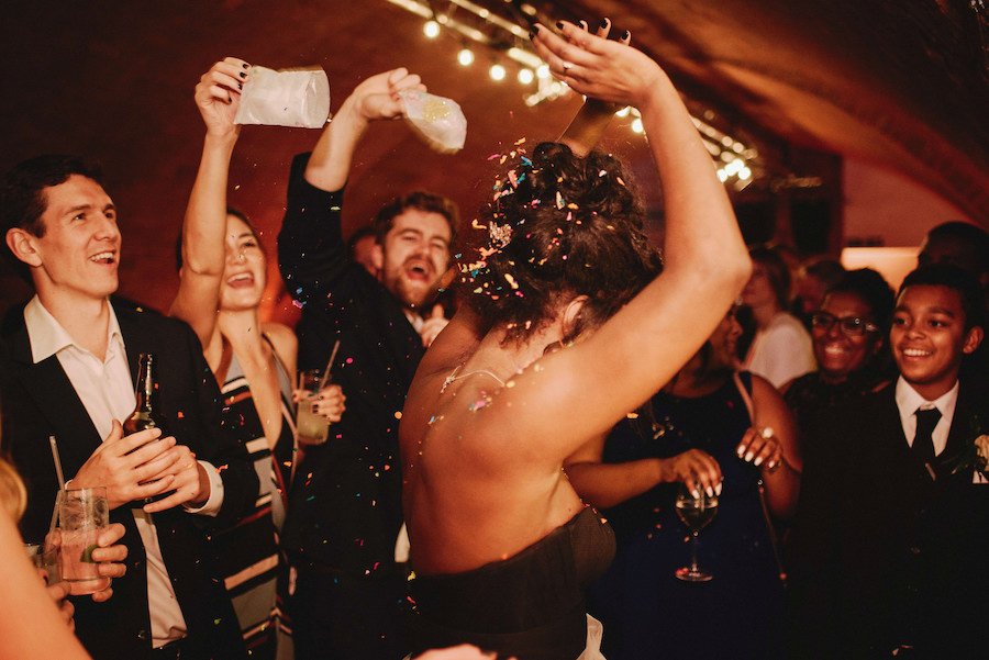 A woman dances in the centre of a warmly lit space as her friends and family cheer and toss confetti.