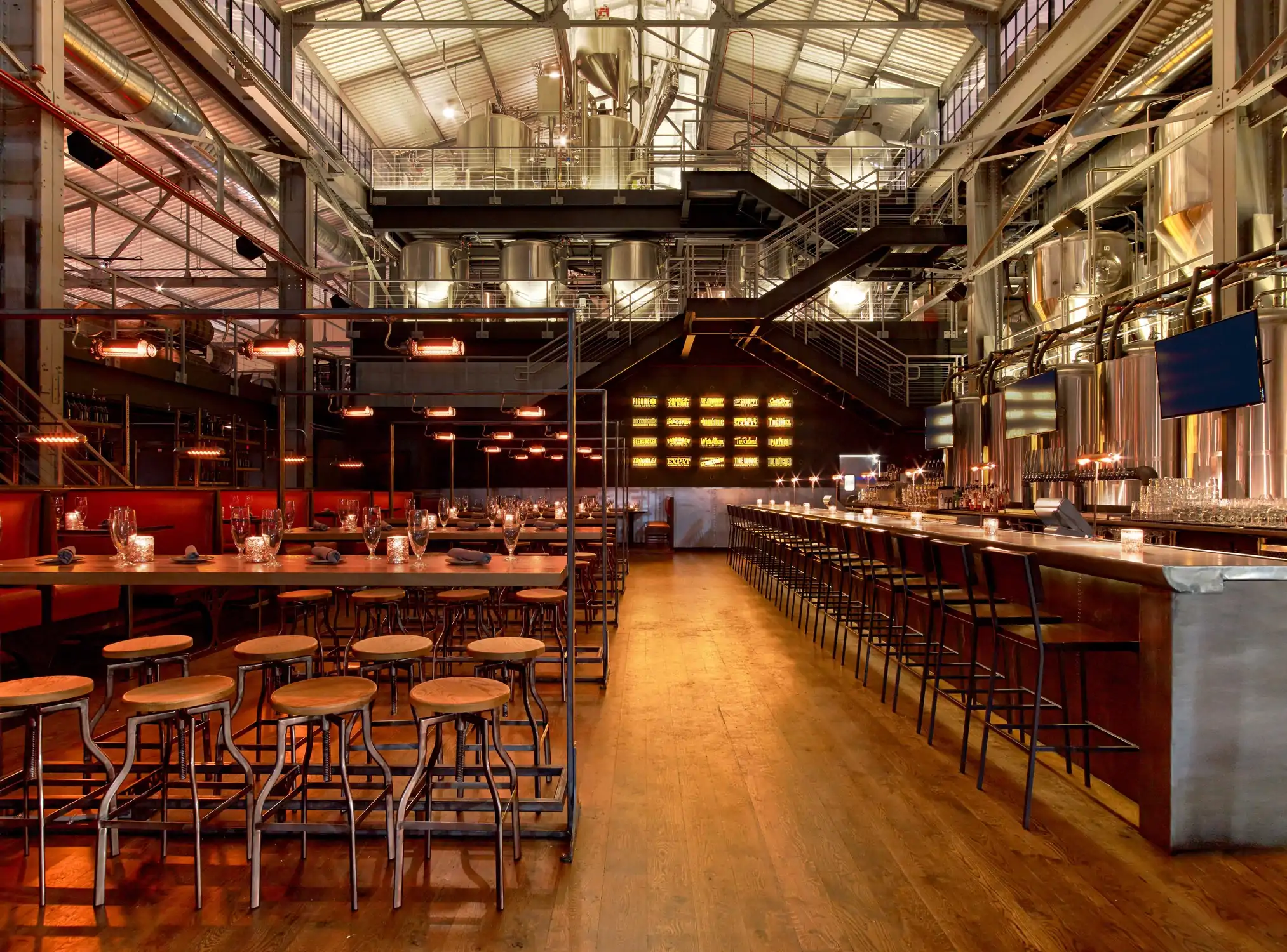 The interior of a brewery with wooden floors and stools, metal tables, shiny brewing equipment and warm lighting.