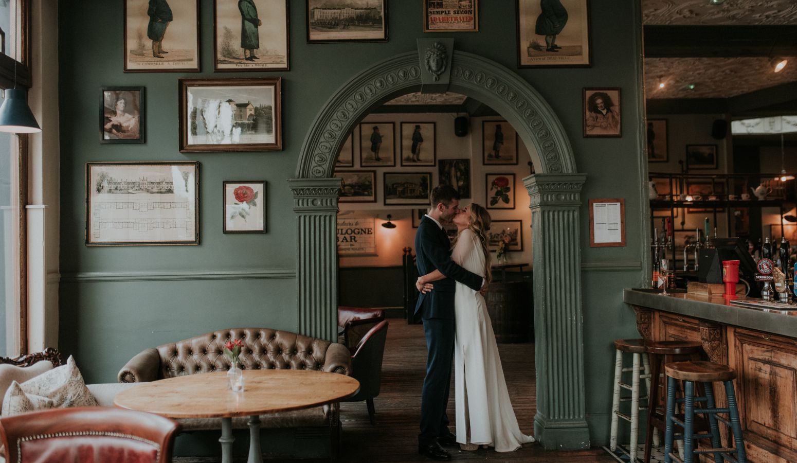A bride and groom embrace in a pub with sage-green walls and wooden furnishings.