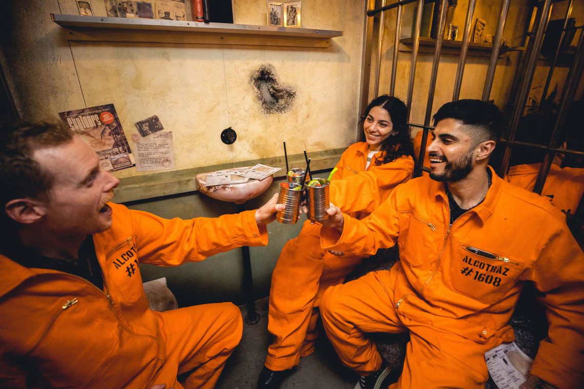 A man and a woman in orange prison jumpsuits sit behind bars, talking and laughing over martinis.