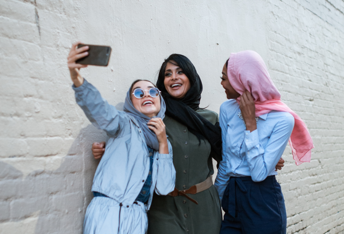 Three girls in head scarfs smile for a selfie in front of a white brick wall.