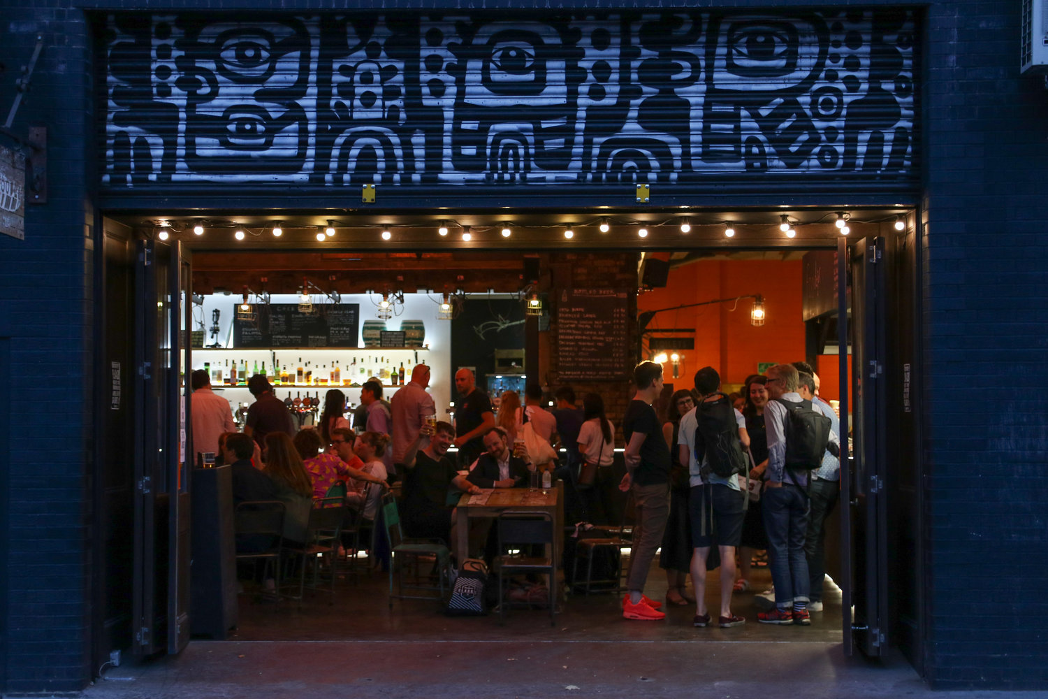 People pack into the warehouse-like space of Doodle Bar, visible under warm lights and a blue metal awning.