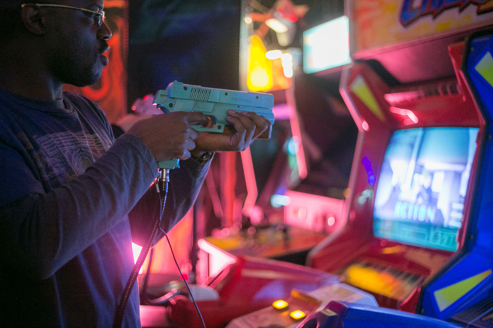 In pink-tinted arcade lighting at Four Quarters, Peckham, a man holds a plastic gun up to the screen of his game.