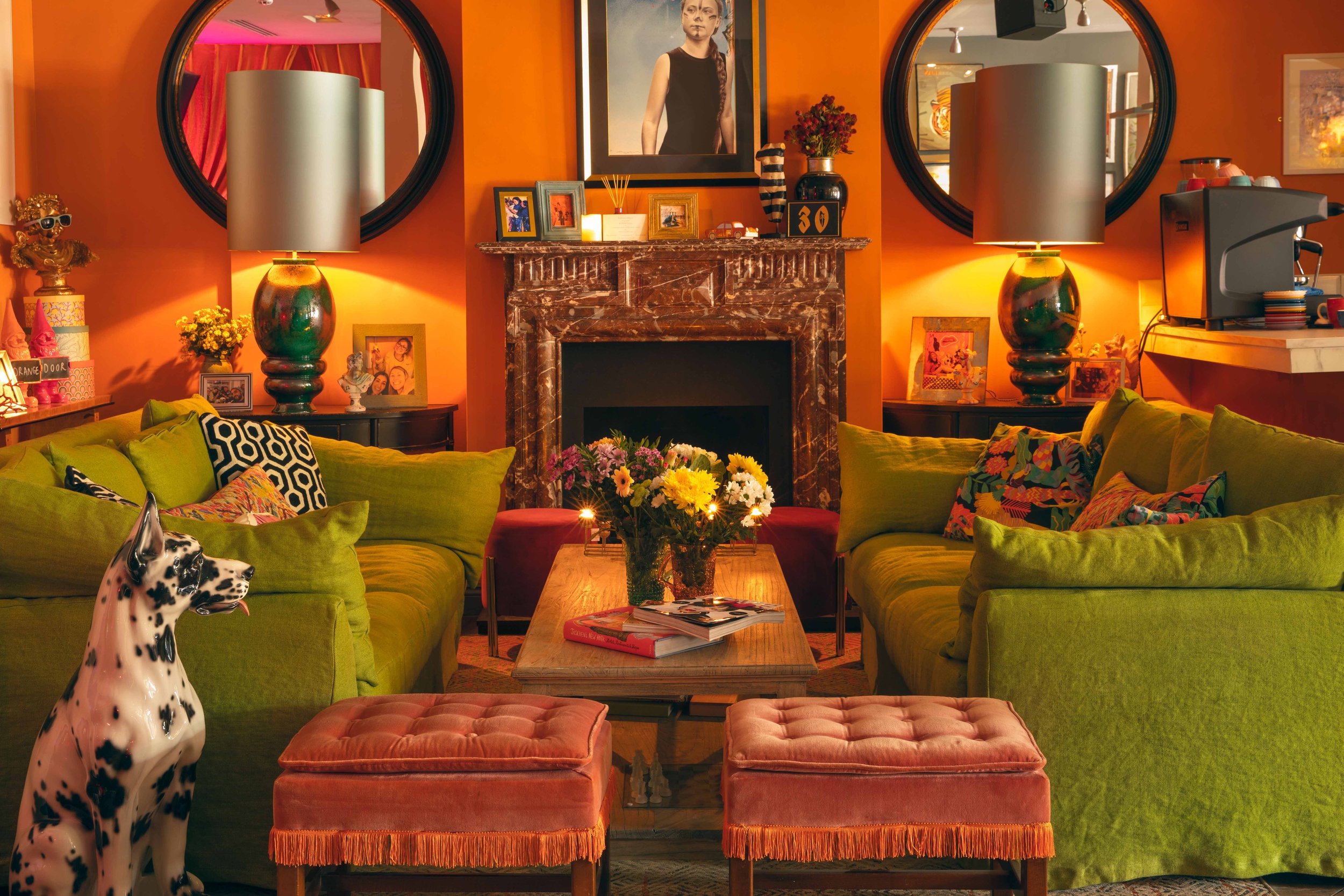 An ornate living room with orange walls, chartreuse couches, and square pink ottomans, plus small tables covered in flowers and picture frames.