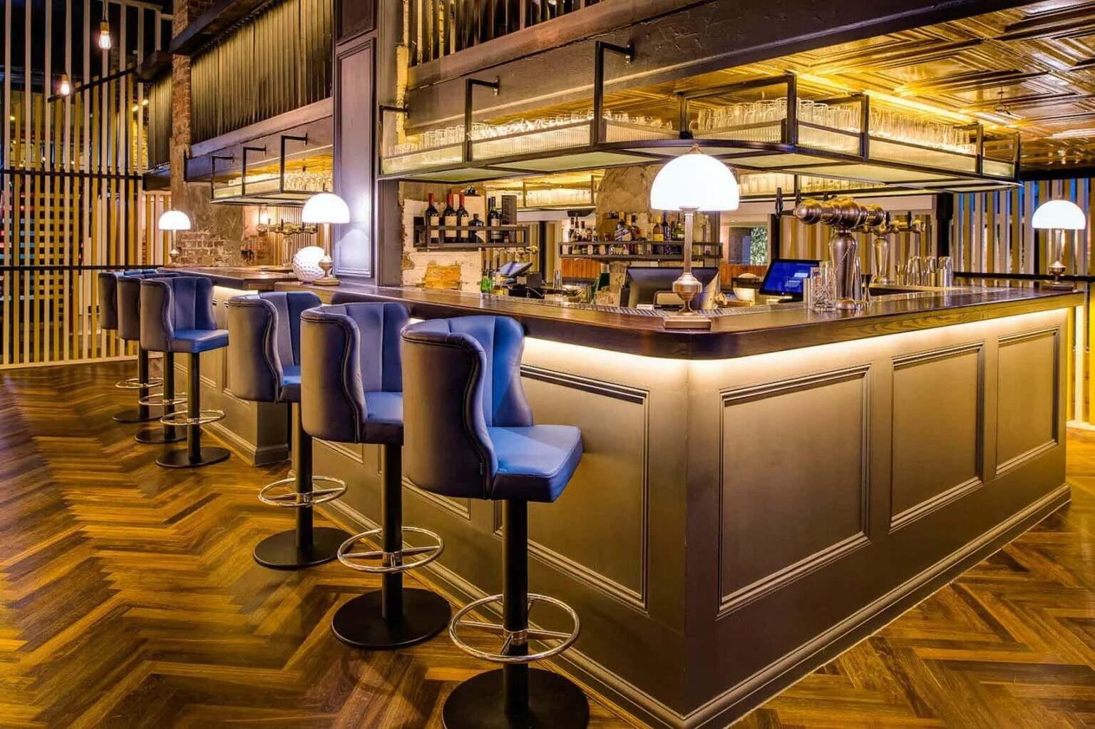 In a gold-lit room with shiny wooden floors, blue barstool chairs are lined up alongside a well-stocked bar.