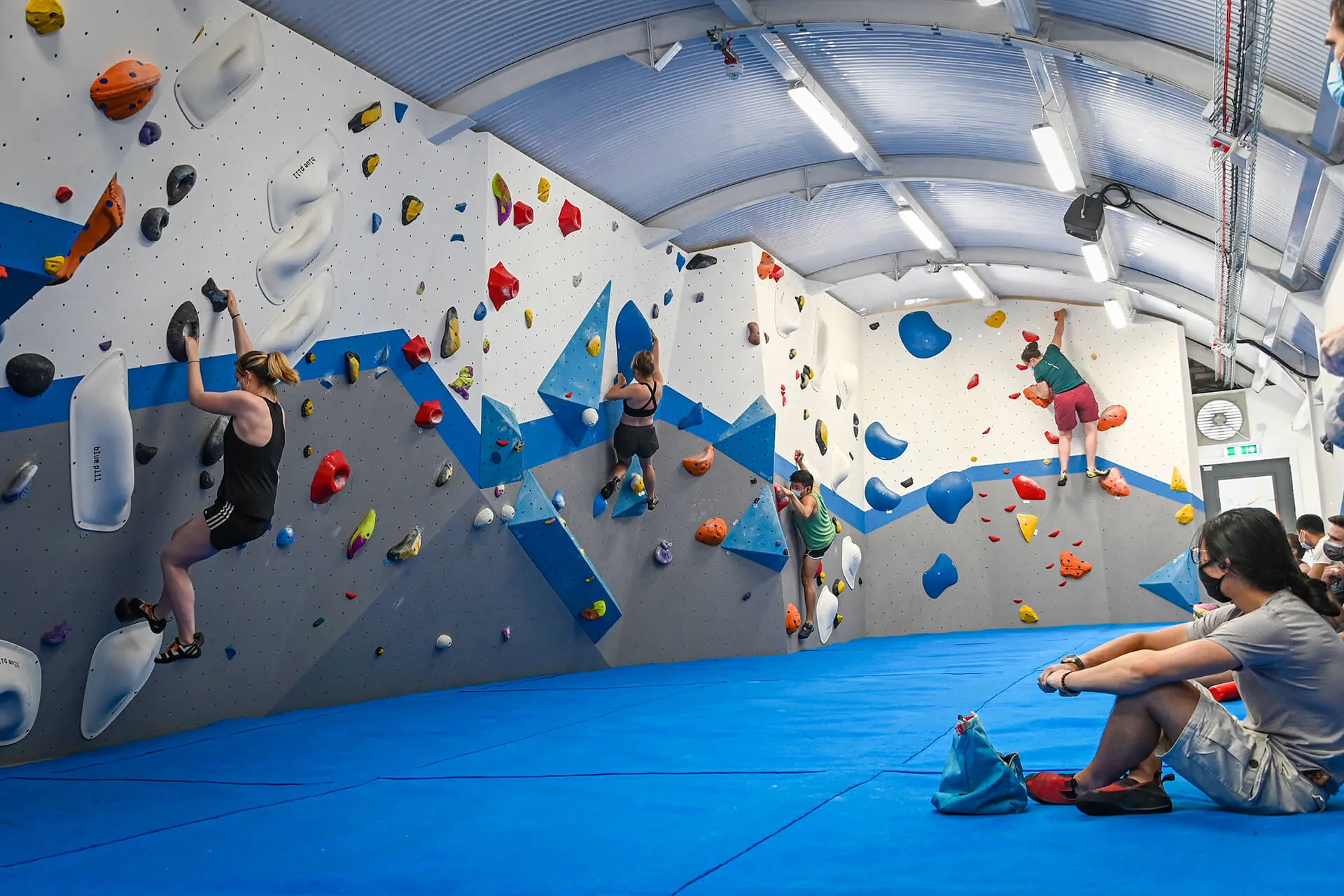 Four climbers move along a climbing wall. A black-haired person watches from the side-line.