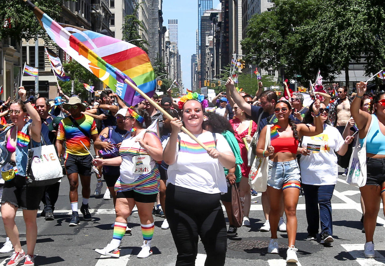 People marching in the NYC Pride Parade, with a woman smiling in the front and waving a rainbow flag.