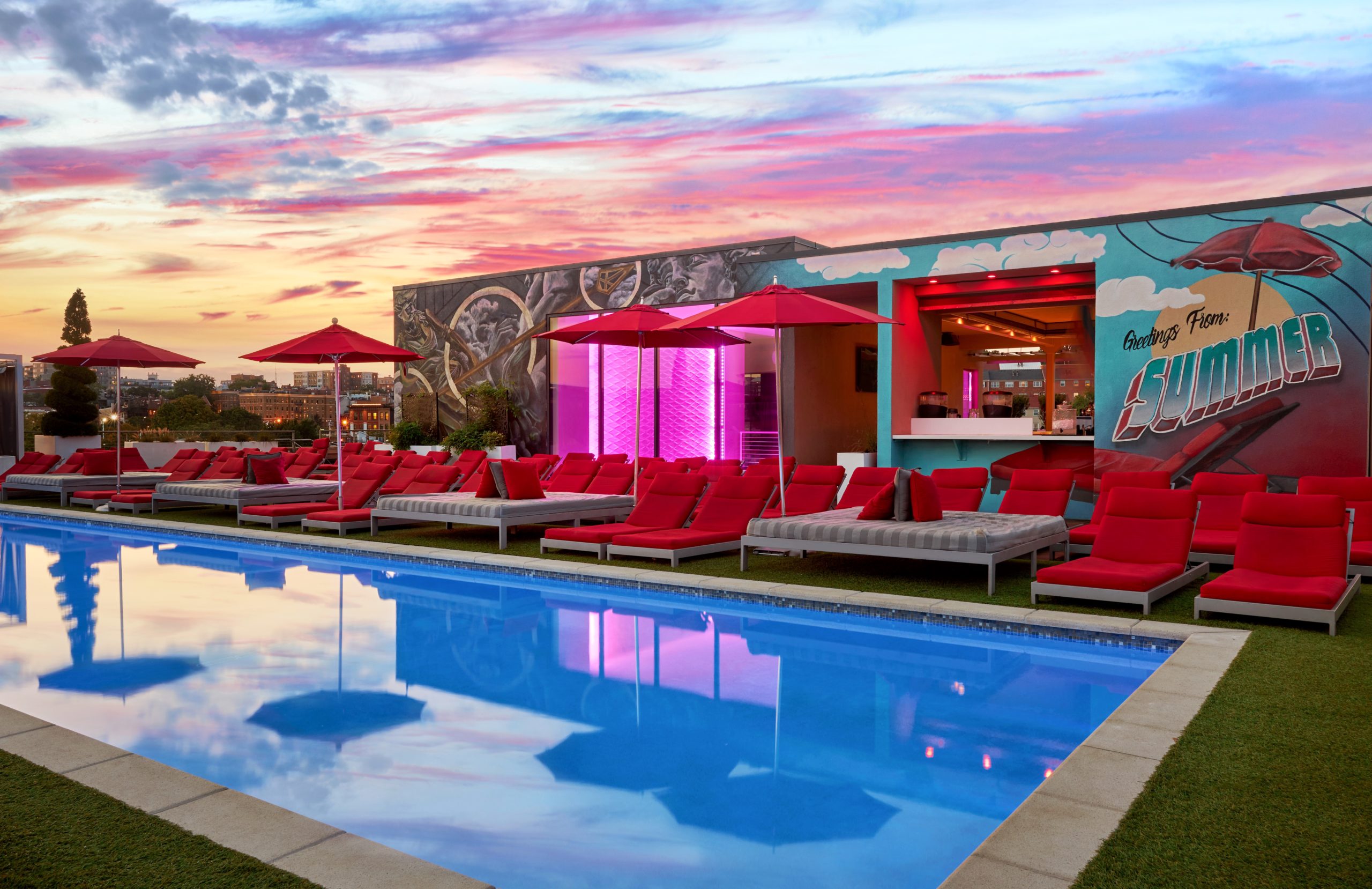 A rooftop pool with a beautiful pink sunset behind it, as well as red lounge chairs and umbrellas on the sides.