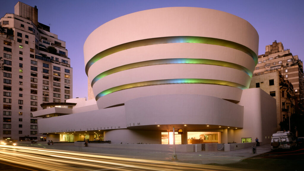 Solomon R. Guggenheim Museum in NYC at dusk, featuring its iconic spiral design with colorful illuminated windows, and adjacent high-rise buildings. 